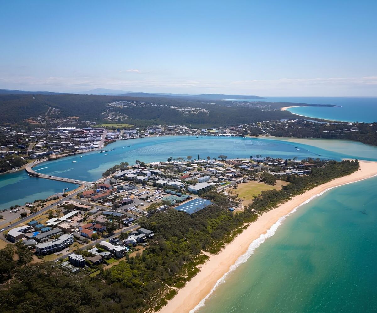 Aerial view of Black Dolphin Holiday Park and Motel in Merimbula, showing the motel buildings, landscaped grounds and nearby Boggy Creek, with its turquoise waters and boat ramp on the NSW Sapphire Coast.