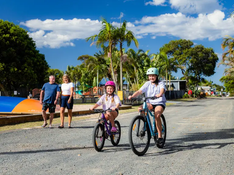 Children riding bikes around a family-friendly holiday park while their family walks nearby.